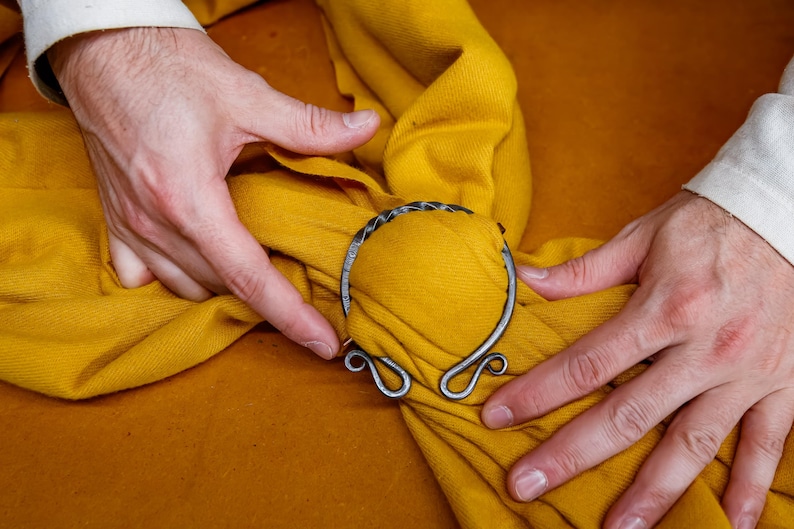 May include: A silver-colored metal cloak clasp with a woven design, set against a mustard yellow fabric. The clasp has a circular shape with decorative swirls. The fabric is being held by hands.