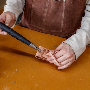 May include: A black dagger with silver detailing is held above a small, tan leather sheath. The dagger has a textured handle and ornate detailing. The sheath is laced with a tan cord. The items are on a wooden surface.