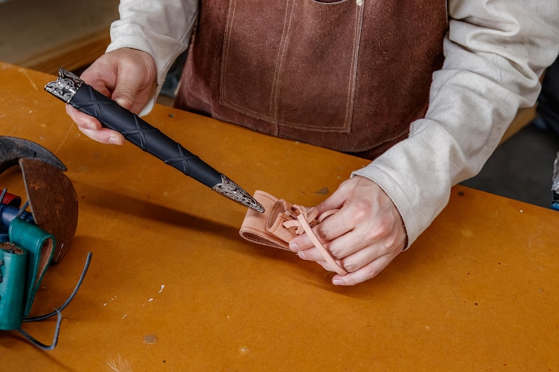 May include: A black dagger with silver detailing is held above a small, tan leather sheath. The dagger has a textured handle and ornate detailing. The sheath is laced with a tan cord. The items are on a wooden surface.