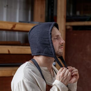 May include: A navy blue quilted hood with ties, worn by a person. The hood is made of a soft, padded material. The person is wearing a light-colored shirt and an apron. The background includes wooden shelves and a brown wall.