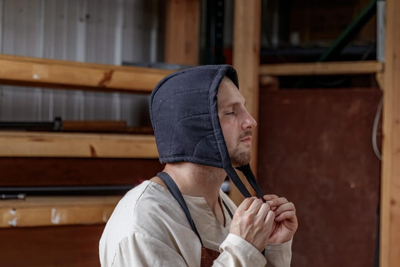 May include: A navy blue quilted hood with ties, worn by a person. The hood is made of a soft, padded material. The person is wearing a light-colored shirt and an apron. The background includes wooden shelves and a brown wall.