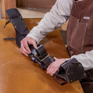 May include: A brown leather tool belt with multiple loops and a metal buckle. The belt is being held by a person's hands.