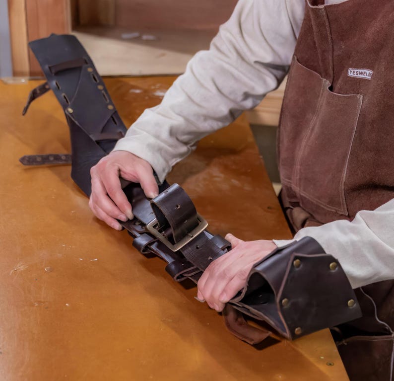 May include: A brown leather tool belt with multiple loops and a metal buckle. The belt is being held by a person's hands.