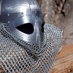 May include: A silver Viking helmet with a riveted brow and a nasal guard. The helmet is paired with a chainmail coif, creating a medieval warrior aesthetic. The metal has a hammered texture, suggesting a handcrafted origin.