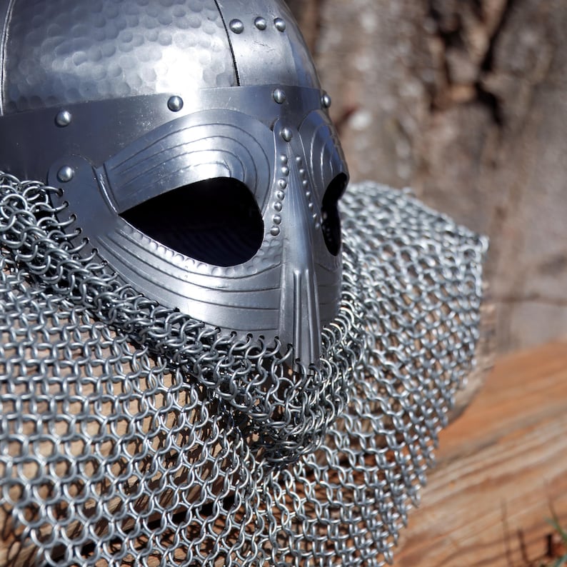 May include: A silver Viking helmet with a riveted brow and a nasal guard. The helmet is paired with a chainmail coif, creating a medieval warrior aesthetic. The metal has a hammered texture, suggesting a handcrafted origin.