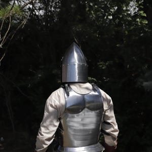 Pu&ograve; includere: Un elmo a bacinetto in acciaio lucido con corona appuntita e una corazza in acciaio abbinata indossata sopra una camicia beige a maniche lunghe. L'armatura presenta superfici metalliche lisce e riflettenti con cinghie in pelle visibili.
