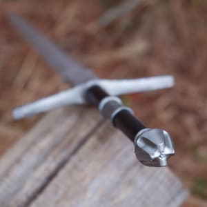 May include: A medieval-style sword with a silver blade and crossguard. The handle is black with silver accents and a silver pommel. The sword is resting on a wooden surface.