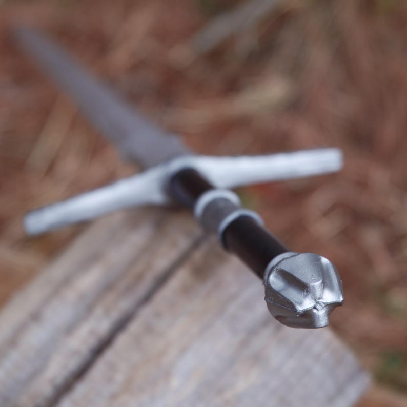 May include: A medieval-style sword with a silver blade and crossguard. The handle is black with silver accents and a silver pommel. The sword is resting on a wooden surface.