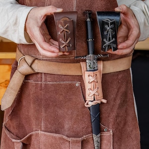 May include: A medieval sword with a black handle and silver detailing, presented with two leather sheaths and a light brown leather scabbard. The sword is held in front of a brown leather apron.