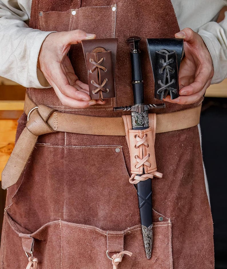 May include: A medieval sword with a black handle and silver detailing, presented with two leather sheaths and a light brown leather scabbard. The sword is held in front of a brown leather apron.