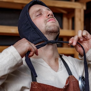 May include: A person wearing a black quilted hood with ties, a light-colored shirt, and a brown apron. The hood is being adjusted with the ties. The background includes wooden shelves.