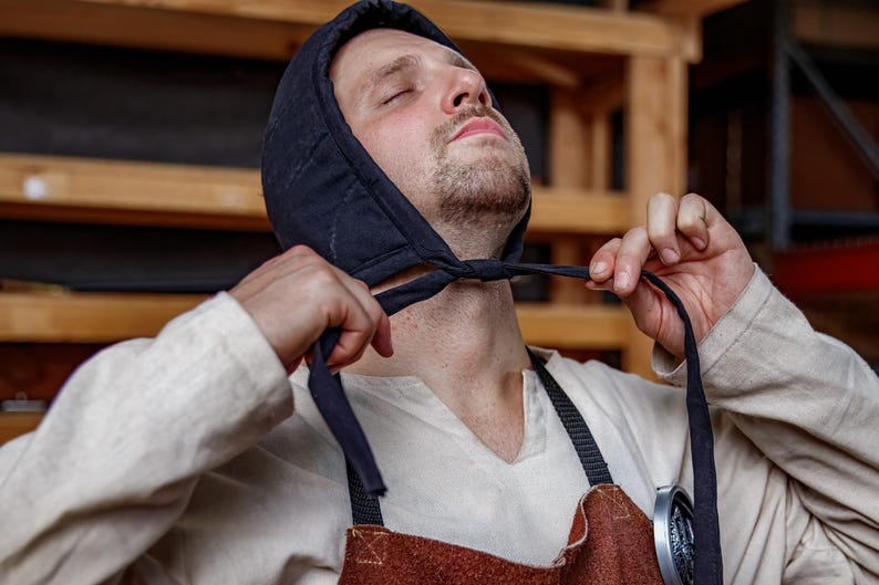 May include: A person wearing a black quilted hood with ties, a light-colored shirt, and a brown apron. The hood is being adjusted with the ties. The background includes wooden shelves.