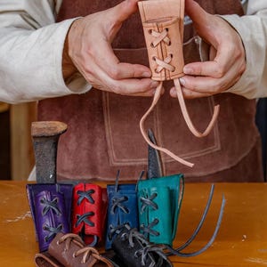 May include: A selection of handcrafted leather sheaths in diverse colours, such as natural tan, purple, red, blue, teal, brown, and black. Each sheath has a laced design and is shown on a wooden surface.