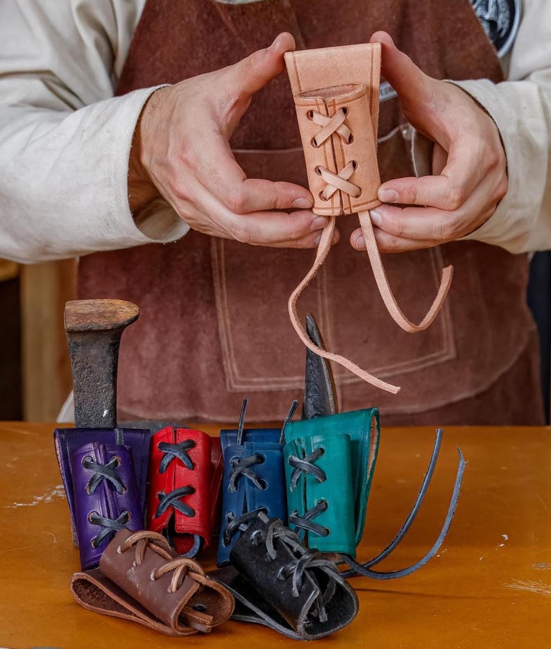 May include: A selection of handcrafted leather sheaths in diverse colours, such as natural tan, purple, red, blue, teal, brown, and black. Each sheath has a laced design and is shown on a wooden surface.