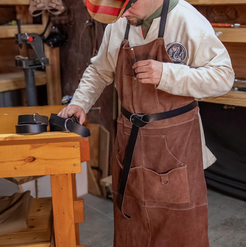 May include: A person wearing a brown leather apron with a black leather belt and a patch on the front. The person is holding two black leather belts in their hands.