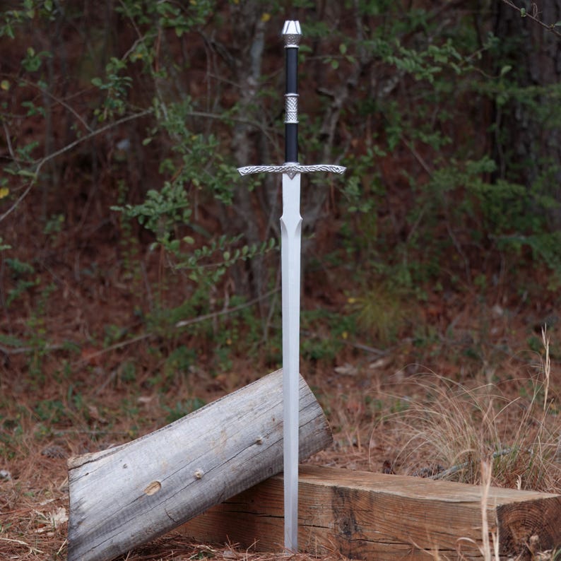 May include: A silver sword with a black handle and ornate silver crossguard. The sword is standing upright, with the blade pointing upwards. The background features a natural setting with trees and foliage.