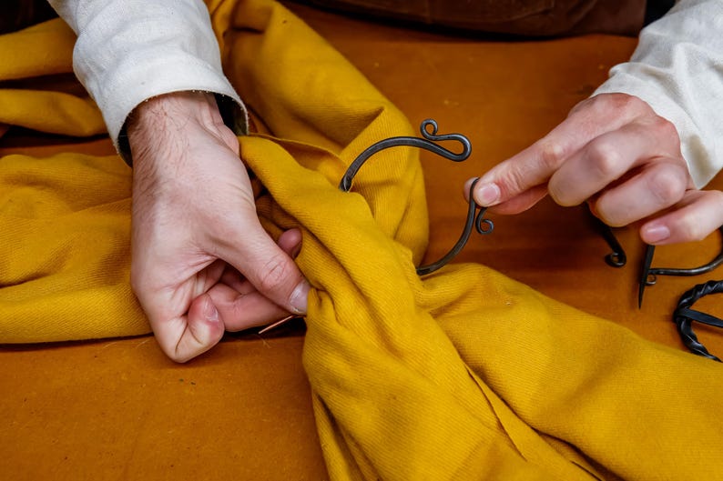 May include: A person's hands are shown working with a piece of yellow fabric and a decorative metal object. The metal object has a dark finish and a curved design with decorative swirls. The fabric is a solid yellow color.