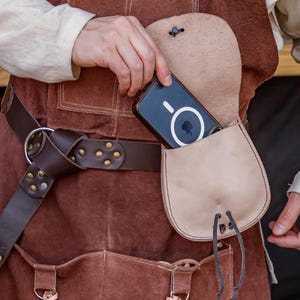 Puede incluir: Un cintur&oacute;n de cuero marr&oacute;n con una hebilla plateada y una bolsa de cuero marr&oacute;n con un tel&eacute;fono negro en su interior. La bolsa est&aacute; unida al cintur&oacute;n con una correa de cuero marr&oacute;n.
