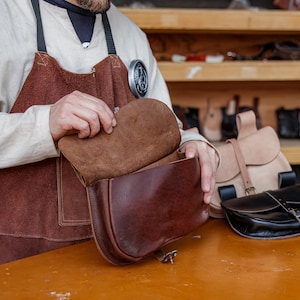Op de afbeelding: Een leerbewerker bekijkt een bruine leren schoudertas met een klep. De tas heeft een bruine su&egrave;de voering. De tas ligt op een houten tafel.