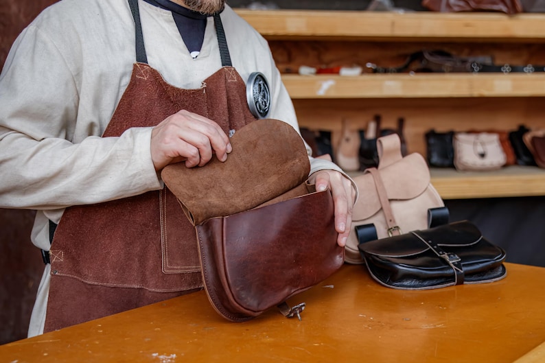 Op de afbeelding: Een leerbewerker bekijkt een bruine leren schoudertas met een klep. De tas heeft een bruine su&egrave;de voering. De tas ligt op een houten tafel.