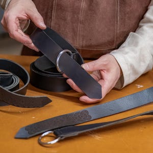 May include: A close-up of a person holding a black leather belt with a silver buckle. The belt is being held over a table with other leather belts.