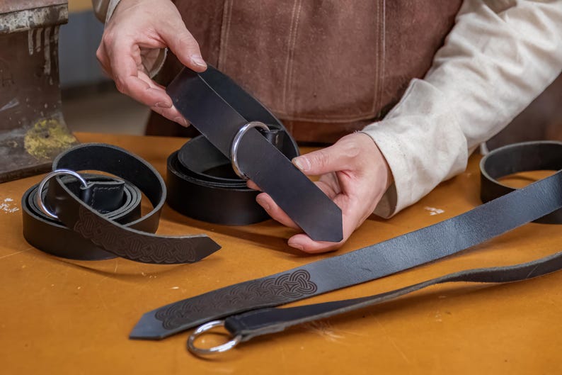 May include: A close-up of a person holding a black leather belt with a silver buckle. The belt is being held over a table with other leather belts.