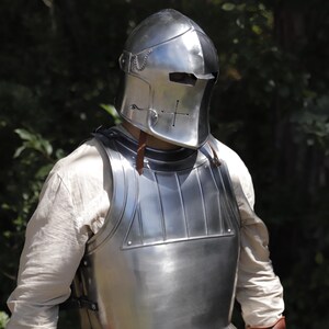 Pu&ograve; includere: Una corazza in acciaio lucido e un elmo chiuso abbinato con una fessura orizzontale per gli occhi e un foro di ventilazione a forma di croce. L'armatura presenta una superficie liscia e riflettente con creste verticali sulla parte superiore.