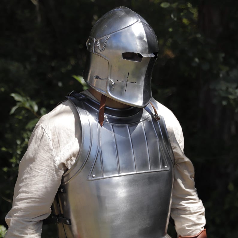 Pu&ograve; includere: Una corazza in acciaio lucido e un elmo chiuso abbinato con una fessura orizzontale per gli occhi e un foro di ventilazione a forma di croce. L'armatura presenta una superficie liscia e riflettente con creste verticali sulla parte superiore.