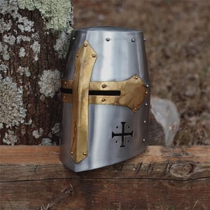 May include: A silver medieval crusader helmet with brass detailing. The helmet has a cross design and is riveted. It is displayed on a wooden surface, with a tree trunk in the background. A historical item.
