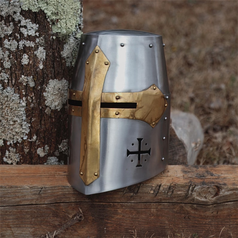May include: A silver medieval crusader helmet with brass detailing. The helmet has a cross design and is riveted. It is displayed on a wooden surface, with a tree trunk in the background. A historical item.