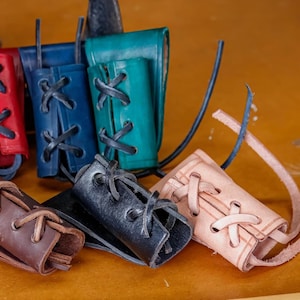 May include: A selection of leather bracers in shades of purple, red, blue, and teal. Each bracer has a laced design with black leather cord. The bracers are displayed on a wooden surface.