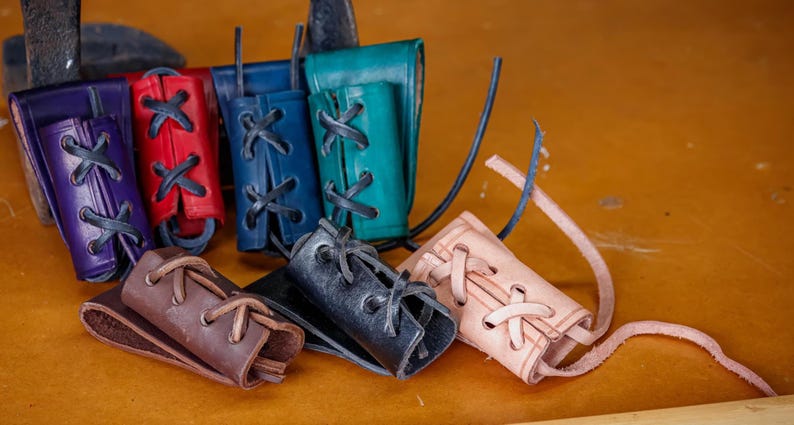 May include: A selection of leather bracers in shades of purple, red, blue, and teal. Each bracer has a laced design with black leather cord. The bracers are displayed on a wooden surface.