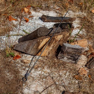 May include: A long, black metal fire poker with a pointed end and a looped handle. The poker is resting on the ground near a pile of firewood. The wood is weathered and shows signs of use, with a mix of light and dark brown tones.