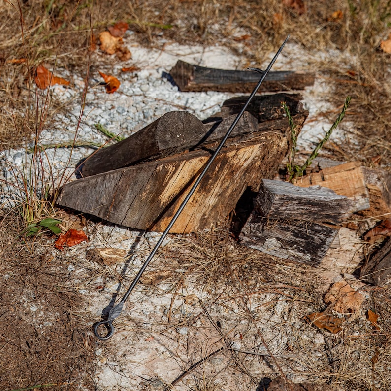 May include: A long, black metal fire poker with a pointed end and a looped handle. The poker is resting on the ground near a pile of firewood. The wood is weathered and shows signs of use, with a mix of light and dark brown tones.