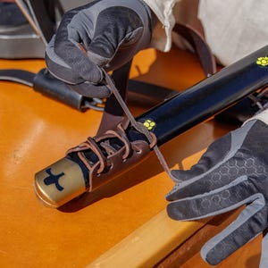 May include: A person is wrapping a brown leather strap around a black sword with a gold handle. The sword has a yellow flower design on the blade. The person is wearing black gloves.