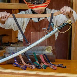 May include: A silver saber with a gold handle and black grip is held up, with a brown leather strap attached. Below are several colored leather sword hangers: purple, brown, black, green, pink, and blue.
