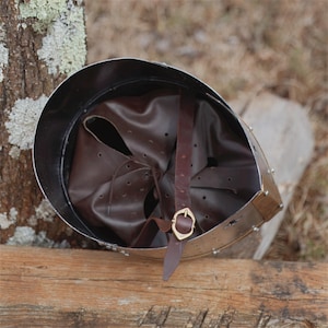 May include: A medieval-style helmet with a polished metal exterior and a brown leather interior. The helmet features a brown leather chin strap with a gold-coloured buckle. The helmet is resting on a wooden surface.