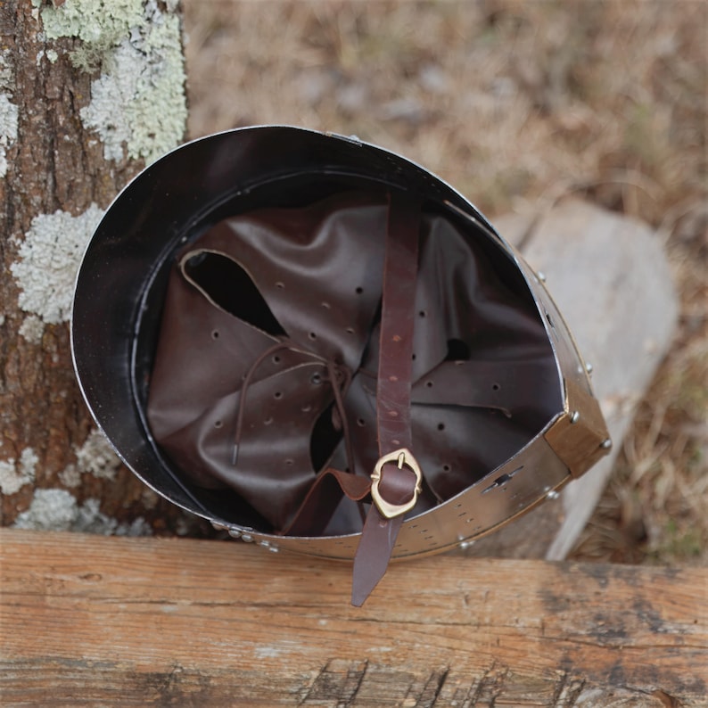 May include: A medieval-style helmet with a polished metal exterior and a brown leather interior. The helmet features a brown leather chin strap with a gold-coloured buckle. The helmet is resting on a wooden surface.