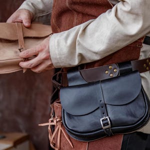 Op de afbeelding: Een persoon draagt een bruin leren vest en een wit shirt en houdt een lichtbruin leren tas vast. De persoon draagt ook een zwarte leren riem met een bruin leren tas eraan.