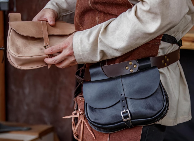 Op de afbeelding: Een persoon draagt een bruin leren vest en een wit shirt en houdt een lichtbruin leren tas vast. De persoon draagt ook een zwarte leren riem met een bruin leren tas eraan.