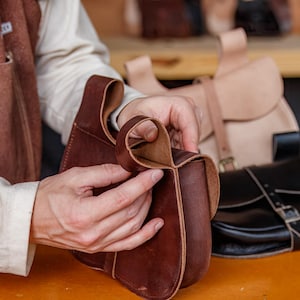 Op de afbeelding: Een leerbewerker werkt aan een bruine leren tas. De tas heeft een klep en een gespsluiting. De leerbewerker houdt de tas in zijn handen en bekijkt hem.