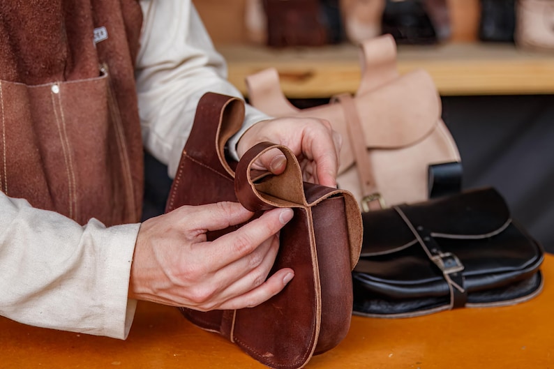 Op de afbeelding: Een leerbewerker werkt aan een bruine leren tas. De tas heeft een klep en een gespsluiting. De leerbewerker houdt de tas in zijn handen en bekijkt hem.