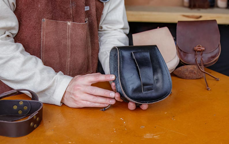 Puede incluir: Una persona sostiene una bolsa de cuero negra con cierre de solapa. La bolsa se sostiene frente a una superficie de madera con otras bolsas de cuero.