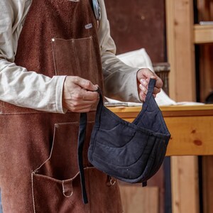 May include: A black quilted oven mitt with a curved design and a long strap. The mitt is held in front of a person wearing a brown leather apron with pockets.