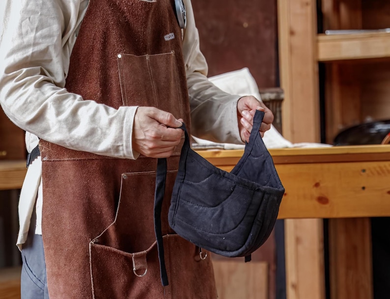 May include: A black quilted oven mitt with a curved design and a long strap. The mitt is held in front of a person wearing a brown leather apron with pockets.