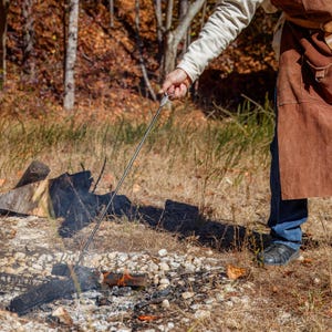 May include: A person using a long, metal roasting stick over a campfire. The stick is held over a small fire with burning wood and embers. The person is wearing a brown apron and blue jeans, with a forest background.