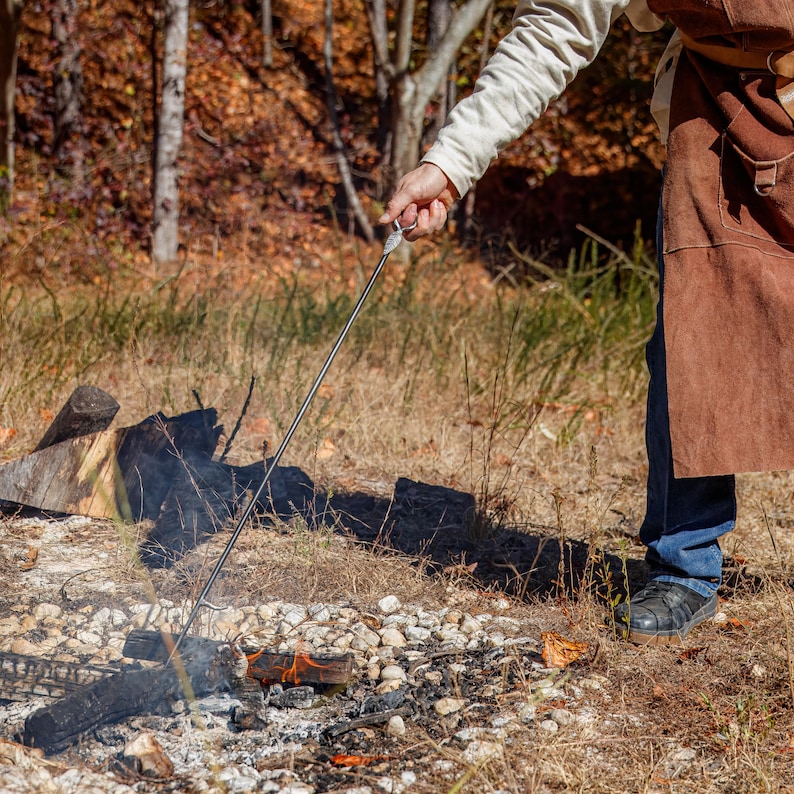 May include: A person using a long, metal roasting stick over a campfire. The stick is held over a small fire with burning wood and embers. The person is wearing a brown apron and blue jeans, with a forest background.