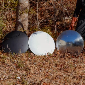 May include: Three large, round metal objects rest on the ground. One is black, one is white, and the third is silver. The silver one reflects the surrounding environment. The objects are outdoors, surrounded by dry leaves and trees.