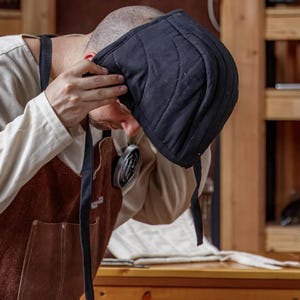 May include: A black quilted cap with a rounded shape and stitched lines. The cap is being held up by a person wearing a tan long-sleeve shirt and a brown apron with black straps. The background includes wooden shelves.