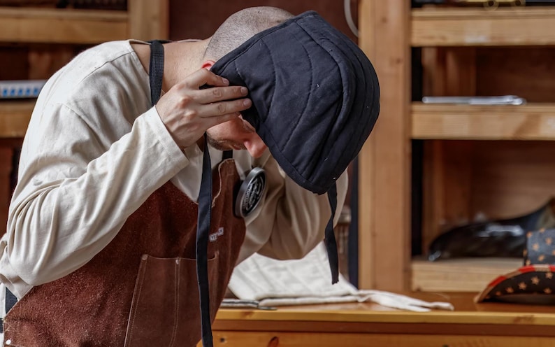 May include: A black quilted cap with a rounded shape and stitched lines. The cap is being held up by a person wearing a tan long-sleeve shirt and a brown apron with black straps. The background includes wooden shelves.